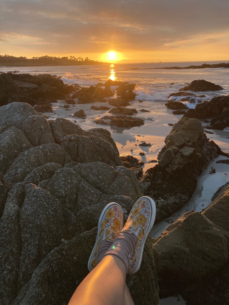 A photograph of the author watching the sunset on the rocks by the Pacific Ocean in California.