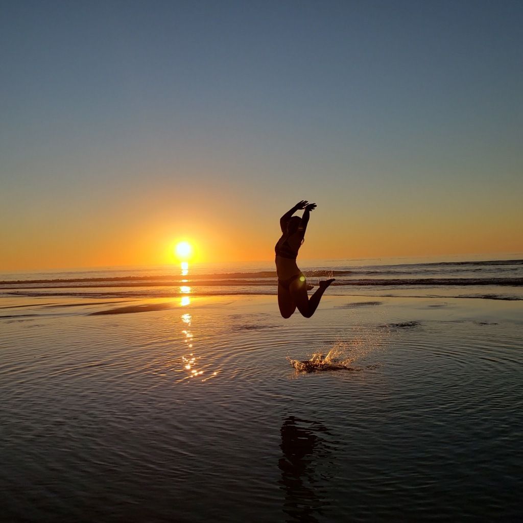 Photograph of a silhouetted girl jumping for joy in the Pacific Ocean with the sun setting behind her. 
