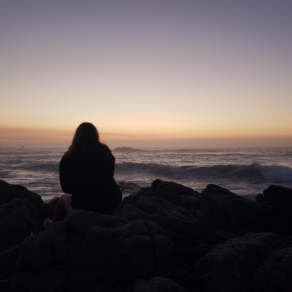 Photograph of a girl silhouetted sitting on rocks in front of the Pacific Ocean in the fading light of sunset.  